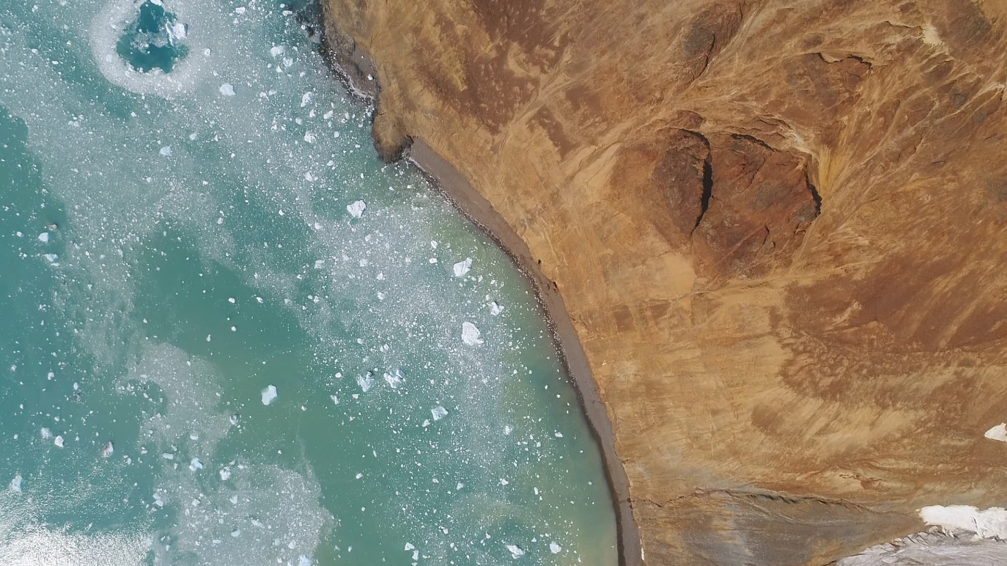 Aerial view of a turquoise lagoon meeting dark volcanic rock on the Antarctic coastline