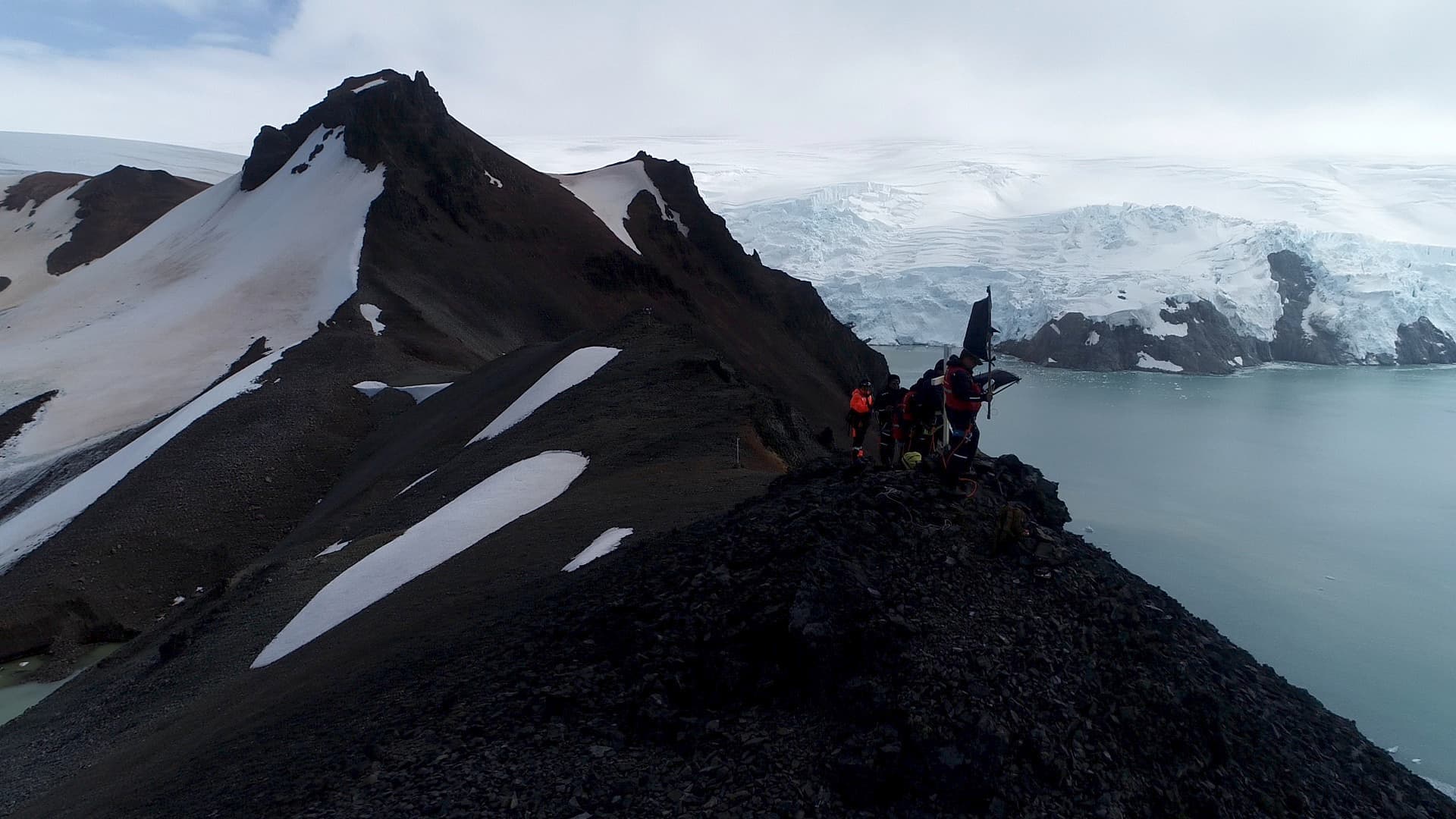 A member of the expedition team holding the mission flag on a ridge, glacier behind, backlit
